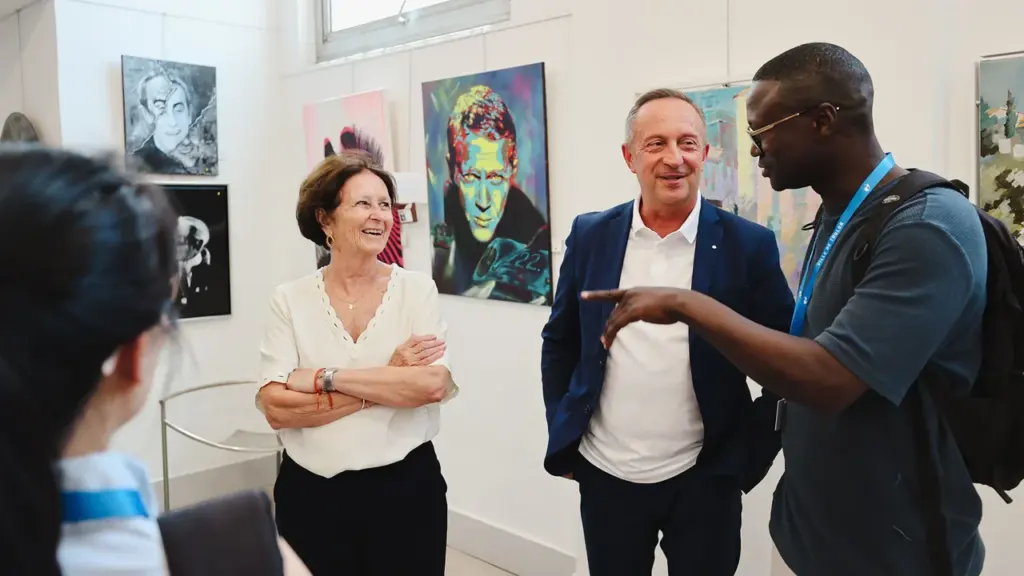 Trois personnes discutent devant des peintures colorées exposées dans une galerie d'art lumineuse.