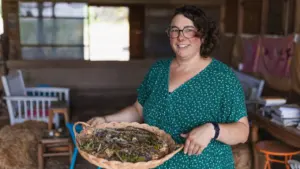 Une femme souriante aux cheveux bruns portant des lunettes présente un plateau tressé d'herbes sauvages.
