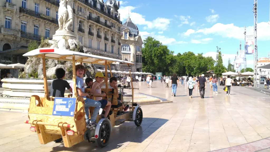 Des enfants souriants circulent en rosalie en bois sur la place de la Comédie.