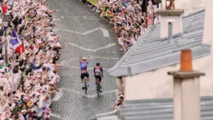 Deux cyclistes professionnels grimpent une rue pavée entourée d'une foule enthousiaste et nombreuse.