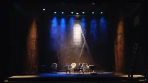 Scène de théâtre avec des chaises éclairées par des projecteurs bleus sur un plateau.
