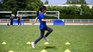 Une jeune fille en t-shirt bleu court sur une pelouse parsemée de cônes jaunes.