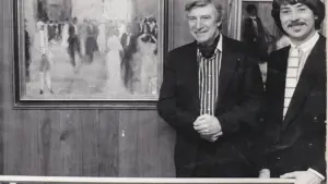 Deux hommes souriants posent devant un tableau représentant une scène de bal en noir et blanc.