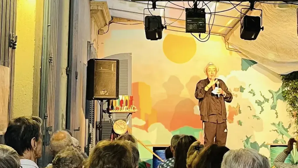 Un homme en chapeau parle devant un public attentif sur une scène intérieure décorée.